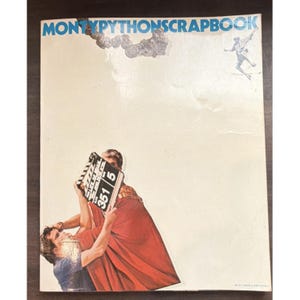 Peut inclure: Un livre à couverture souple intitulé MONTYPYTHONSCRAPBOOK avec une couverture crème. Le haut présente une petite illustration en noir et blanc d'une silhouette et de fumée. En bas, une personne en robe rouge tient un clap de cinéma.