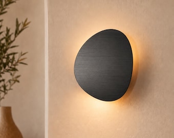 May include: A wall-mounted light fitting with an asymmetrical, pebble-shaped black metal cover. The fitting emits a soft, warm glow from behind its edges, creating a halo effect against the wall surface it is mounted upon.