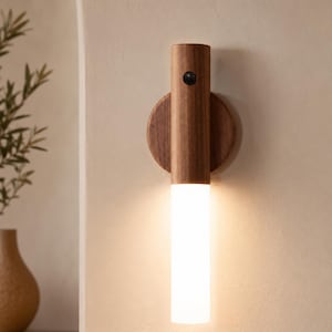 May include: A wall-mounted light fixture featuring a cylindrical dark wood upper section with a small circular sensor and a matching circular wood backplate. The lower portion consists of a frosted white cylindrical light diffuser emitting a warm glow.