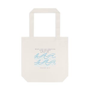 Könnte beinhalten: Eine cremefarbene Stofftasche aus Segeltuch mit zwei Schulterriemen. Auf der Vorderseite befindet sich eine Grafik mit hellblauen Wellen und dem schwarzen Text: WHEN YOU GO THROUGH DEEP WATERS I WILL BE WITH YOU, gefolgt von ISAIAH 43:2.