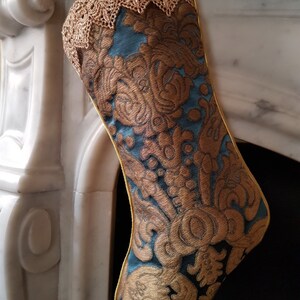 May include: A decorative Christmas stocking with a teal and gold floral pattern. The stocking has a gold lace trim at the top and a gold trim along the edges. The stocking is hanging in front of a white fireplace.