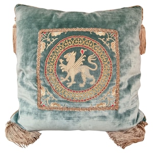 May include: A teal blue velvet cushion with a gold and red embroidered design featuring a mythical creature. The cushion has tassels on each corner.