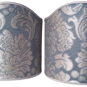May include: Two matching lampshades with a blue-green background and a silver floral pattern. The shades have a curved top and a silver trim along the top and bottom edges. The design is a classic, elegant style.