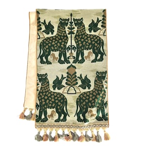 Pode incluir: Um corredor de mesa verde e bege com um padr&atilde;o repetido de leopardos e p&aacute;ssaros estilizados. O corredor tem uma borda de franja com borlas.