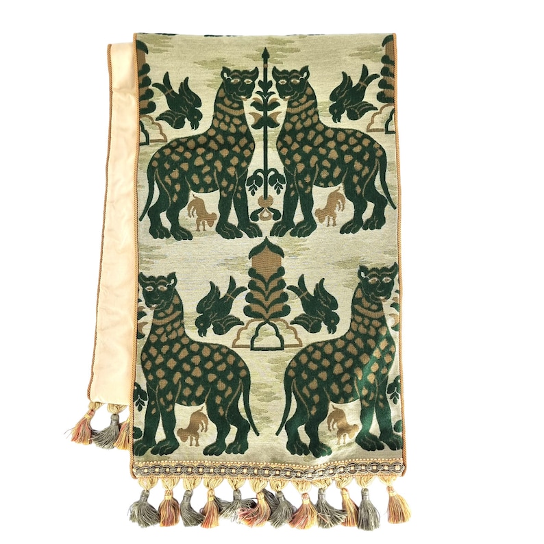 Pode incluir: Um corredor de mesa verde e bege com um padr&atilde;o repetido de leopardos e p&aacute;ssaros estilizados. O corredor tem uma borda de franja com borlas.