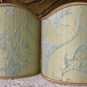 May include: Two light green lampshades with a subtle floral pattern. The lampshades are made of a woven fabric and have a gold trim.