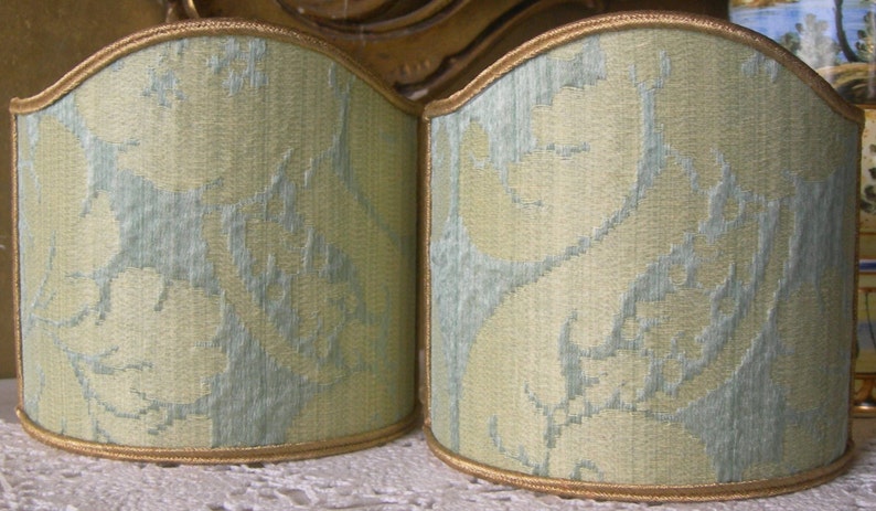 May include: Two light green lampshades with a subtle floral pattern. The lampshades are made of a woven fabric and have a gold trim.