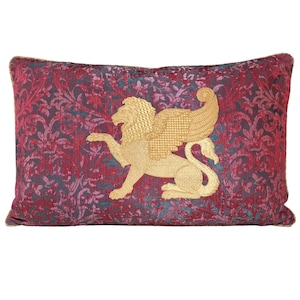 Peut inclure: Coussin rectangulaire décoratif avec un motif floral bordeaux et sarcelle. Un lion ailé brodé en or est centré sur le coussin. Le coussin a une bordure en corde dorée.