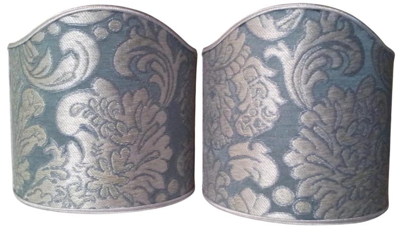 May include: Two matching lampshades with a blue-green background and a silver floral pattern. The shades have a curved top and a silver trim along the top and bottom edges. The design is a classic, elegant style.