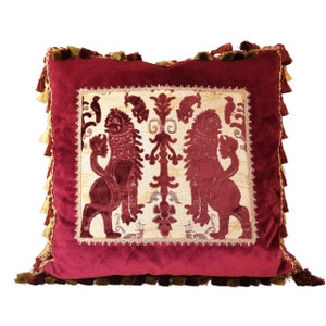 Puede incluir: Una almohada de terciopelo burdeos con un panel bordado decorativo que presenta dos leones y un diseño floral. La almohada tiene un borde con flecos.