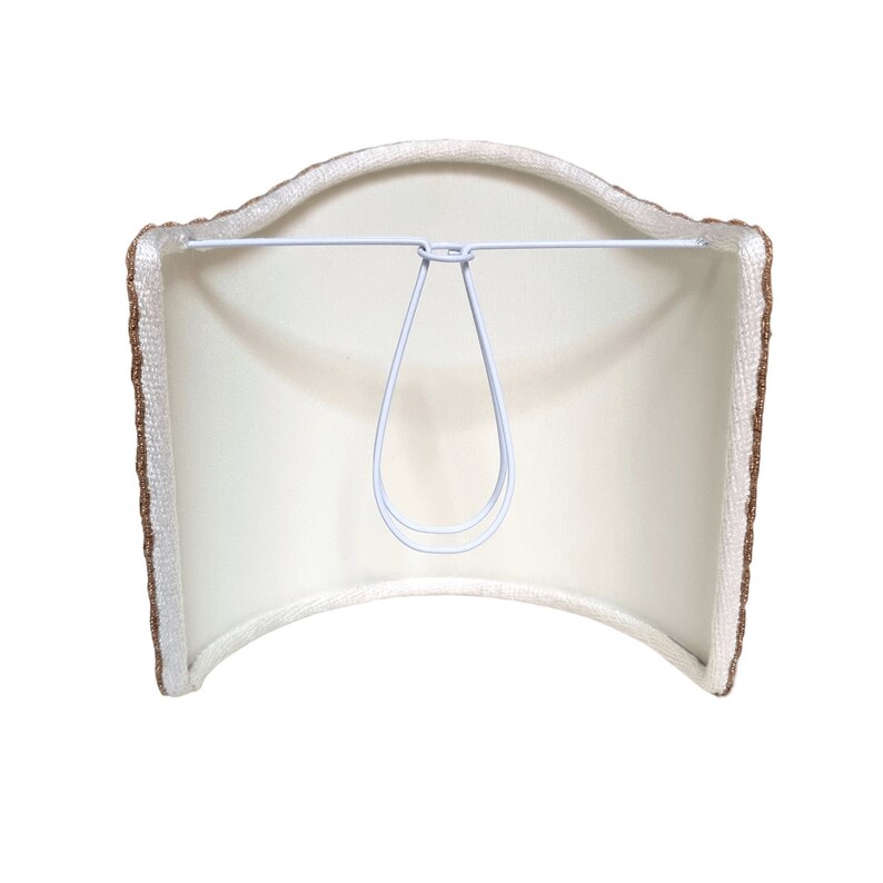 May include: A white lampshade with a brown trim. The shade is curved and has a metal wire loop for hanging.