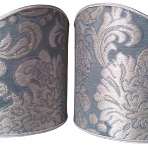 May include: Two decorative lamp shade covers. The shades are a muted teal color with a silver floral damask pattern. The top and bottom edges are trimmed with a silver border. The shades are cylindrical with a curved top.