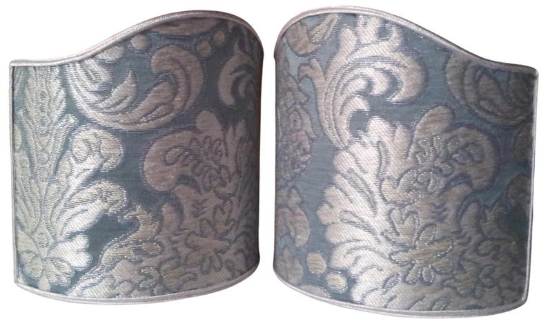 May include: Two decorative lamp shade covers. The shades are a muted teal color with a silver floral damask pattern. The top and bottom edges are trimmed with a silver border. The shades are cylindrical with a curved top.