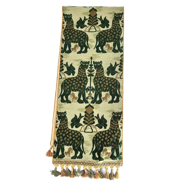 Pode incluir: Um corredor de mesa verde e dourado com um padr&atilde;o repetido de leopardos estilizados. O corredor tem uma borda de franja com borlas douradas e verdes.