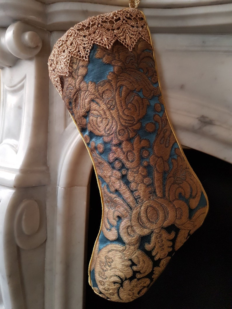May include: A decorative Christmas stocking with a teal and gold floral pattern. The stocking has a gold lace trim at the top and a gold trim along the edges. The stocking is hanging in front of a white fireplace.