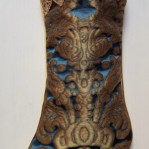 May include: A decorative Christmas stocking with a gold and teal brocade pattern. The stocking features a gold lace trim at the top and a gold hanging loop. The brocade design includes ornate floral and scroll motifs.