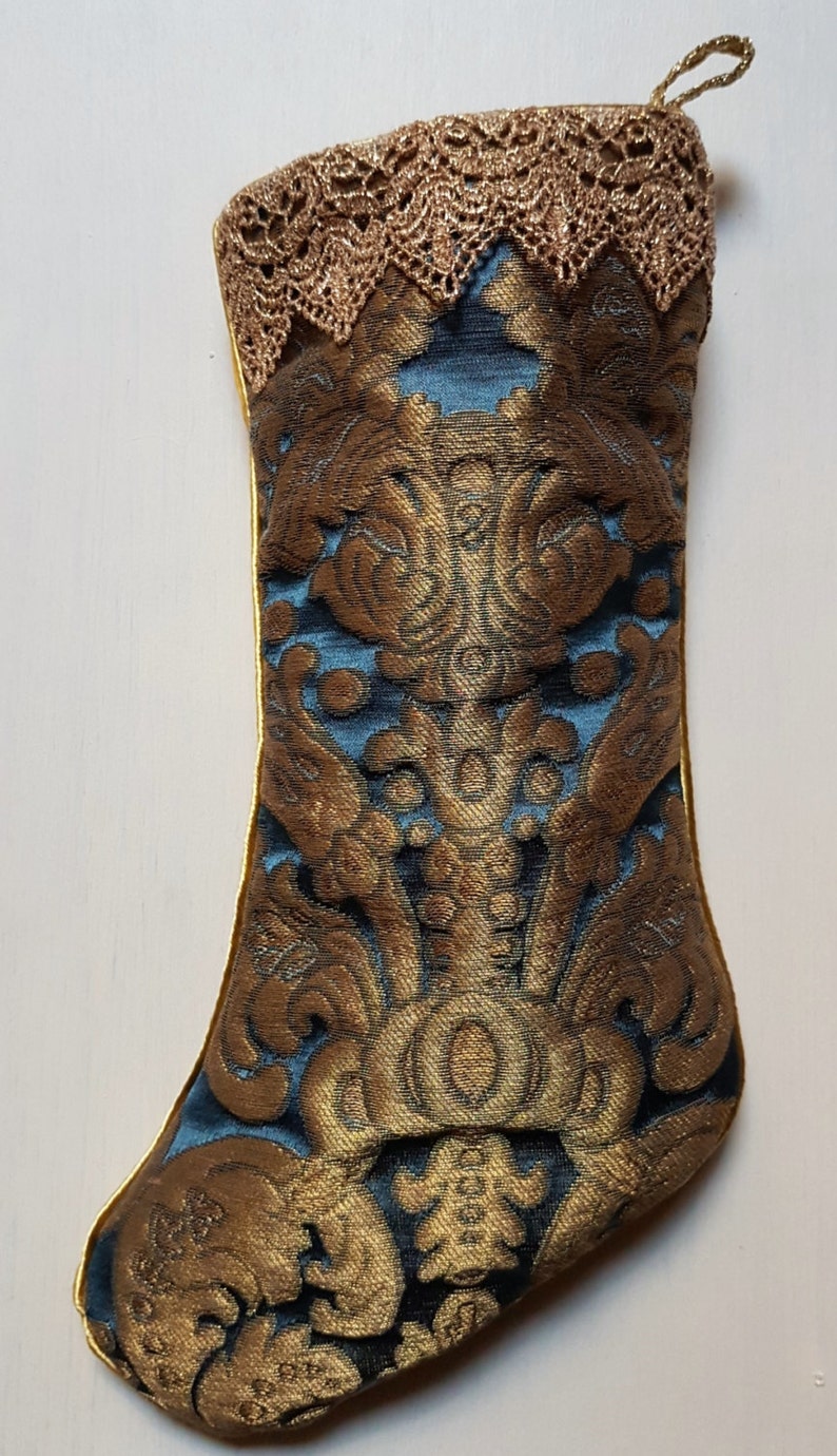 May include: A decorative Christmas stocking with a gold and teal brocade pattern. The stocking features a gold lace trim at the top and a gold hanging loop. The brocade design includes ornate floral and scroll motifs.