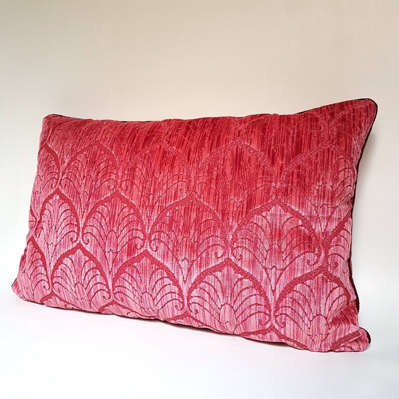 May include: A pink velvet pillow with a decorative pattern of stylized leaves. The pillow has a dark brown border.