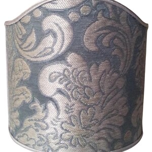 May include: A cylindrical lampshade with a damask pattern in shades of blue and beige. The fabric has a textured appearance, and the top and bottom edges are trimmed with a silver-colored piping.