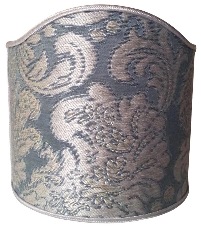 May include: A cylindrical lampshade with a damask pattern in shades of blue and beige. The fabric has a textured appearance, and the top and bottom edges are trimmed with a silver-colored piping.