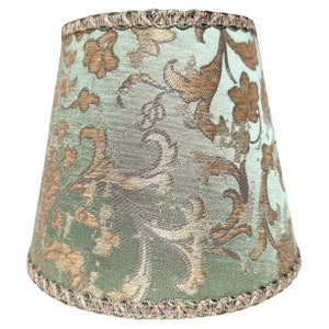 Pode incluir: Um abajur verde com um padr&atilde;o floral dourado e marrom. O abajur tem uma guarni&ccedil;&atilde;o decorativa ao redor da parte superior e inferior.