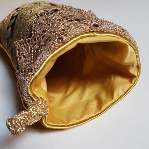 May include: A gold-colored pouch with a drawstring closure. The pouch features a gold lace trim and a gold satin lining. The exterior is a patterned gold fabric with black accents. The pouch is open, revealing the interior.