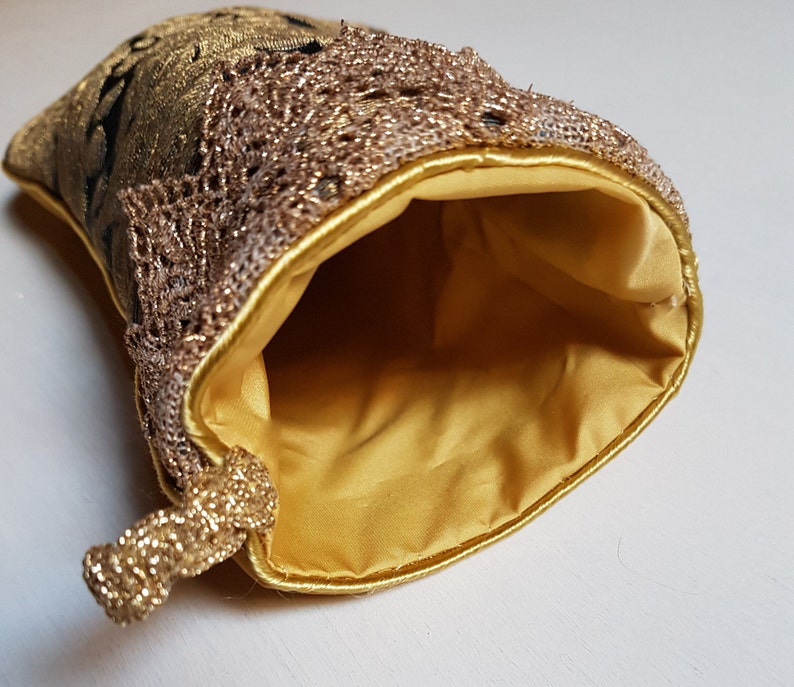 May include: A gold-colored pouch with a drawstring closure. The pouch features a gold lace trim and a gold satin lining. The exterior is a patterned gold fabric with black accents. The pouch is open, revealing the interior.