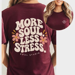 Könnte beinhalten: Ein kastanienbraunes T-Shirt mit dem Text MORE SOUL LESS STRESS in großer, cremefarbener Retro-Schrift auf der Rückseite, akzentuiert durch zwei rosa Blumen. Die Vorderseite zeigt eine kleine weiße Strichzeichnung einer sitzenden Yoga-Figur.