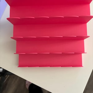 Puede incluir: Un expositor de niveles de color rosa brillante con cuatro estantes horizontales. Cada estante está diseñado con un borde festoneado en la parte frontal, proporcionando una superficie estructurada para organizar o exhibir diversos objetos pequeños.