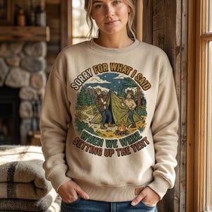 May include: A beige crewneck sweatshirt featuring a graphic illustration of two people setting up a tent in a forest with a campfire. The text reads, "SORRY FOR WHAT I SAID WHEN WE WERE SETTING UP THE TENT" in a curved, retro-style font.