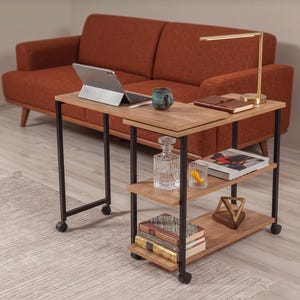 Puede incluir: Una mesa auxiliar móvil con marco de metal negro y estantes de madera clara. La superficie superior sostiene una tableta, una taza, un cuaderno y una lámpara dorada. Los estantes inferiores contienen un decantador, un vaso, una escultura y libros.