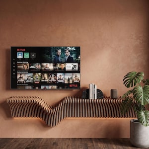 Puede incluir: Un televisor montado en la pared que muestra la interfaz de Netflix, colocado sobre un estante de madera largo y horizontal con listones y un diseño ondulado. En el estante hay sujetalibros de mármol negro, varios libros y un pequeño altavoz cilíndrico.