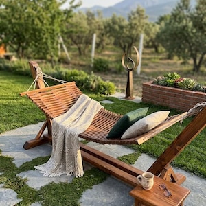 Puede incluir: Una hamaca de madera con diseño de listones, que incluye una manta beige estampada, un cojín verde oscuro y un cojín beige. Una pequeña mesa auxiliar de madera sostiene una taza de cerámica y unas gafas, todo sobre un patio de piedra.
