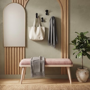 May include: A pink upholstered bench with light wood legs, featuring a grey textured throw blanket. Above, a cream canvas tote bag and a grey scarf hang from three black wall hooks next to an arched mirror. A green potted plant sits nearby.