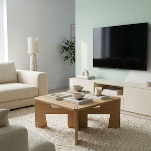 May include: A square light-toned wooden coffee table with a geometric base, featuring a flat top surface holding several stacked books, two small ceramic bowls, and a cylindrical candle, positioned in a living room with a television and sofa.