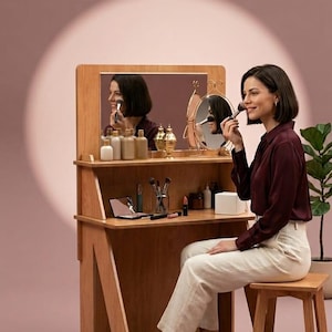 May include: A light brown wooden vanity table with a rectangular mirror, a smaller oval mirror, and a matching wooden stool. The vanity surface holds various cosmetic bottles, makeup brushes in a glass holder, and a small open compact mirror.