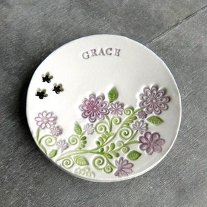 Peut inclure: Plat en céramique blanche avec un motif floral dans les tons de violet et de vert. Le plat porte le mot "GRACE" en relief sur le dessus.