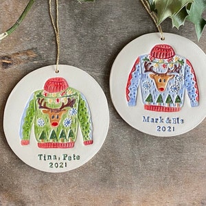 May include: Two round ceramic ornaments with a reindeer wearing a Christmas sweater design. The ornaments are white with a gold string for hanging. The top ornament has a blue sweater with the text "Mark & Els 2021". The bottom ornament has a green sweater with the text "Tina, Pete 2021".