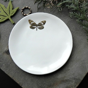 Può includere: Un piatto portaoggetti in ceramica bianca con un design a farfalla dorata. Il piatto è rotondo e ha un bordo leggermente rialzato.