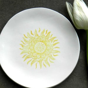May include: A white ceramic plate with a yellow sun mandala design. The plate is on a wooden surface.