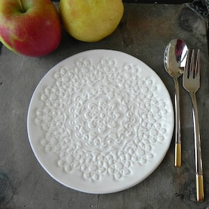 Pode incluir: Prato de cer&acirc;mica branco com um design de renda em relevo. O prato est&aacute; vazio e tem um brilho sutil. Dois talheres de prata est&atilde;o sobre uma superf&iacute;cie escura ao lado do prato.