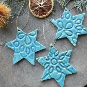 Op de afbeelding: Drie turquoise keramische sterornamenten met reliëf bloempatronen. Elke ster heeft een klein gaatje om op te hangen met een gouden draad. Een gedroogde sinaasappelschijf en dennentakken op de achtergrond.