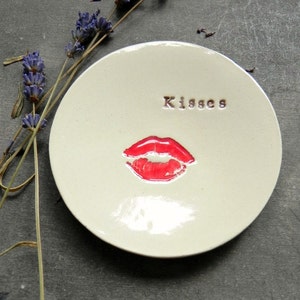 以下が含まれることがあります： 赤い口紅のキスプリントと、茶色の文字で「Kisses」と書かれた白いセラミック製の皿です。