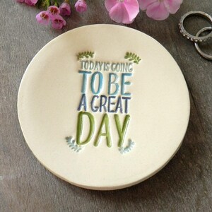 Puede incluir: Un plato de anillo de cerámica blanca con el texto "TODAY IS GOING TO BE A GREAT DAY" en letras azules y verdes. El plato está decorado con hojas verdes y está sobre una superficie de madera.