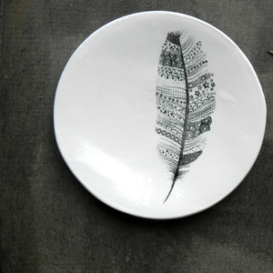 Könnte beinhalten: Ein weißer Keramikteller mit einem schwarz-weißen Federdesign. Die Feder ist mit verschiedenen Mustern und Formen verziert.