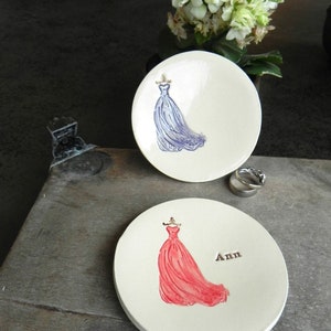 May include: Two ceramic ring dishes with a white background. One dish features a blue dress and the other features a red dress. The red dish has the name "Ann" written on it.