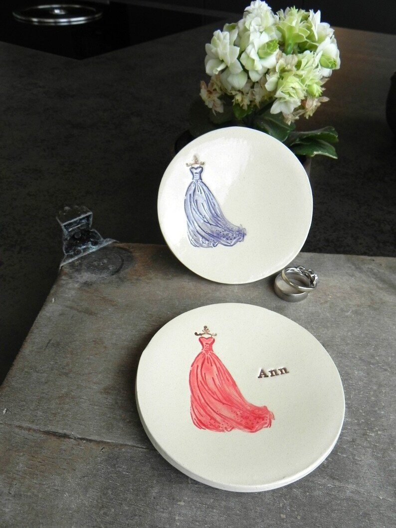 May include: Two ceramic ring dishes with a white background. One dish features a blue dress and the other features a red dress. The red dish has the name "Ann" written on it.