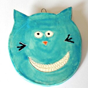 Peut inclure: Une décoration murale en céramique en forme de visage de chat turquoise avec un large sourire aux dents, des moustaches noires et de grands yeux blancs.