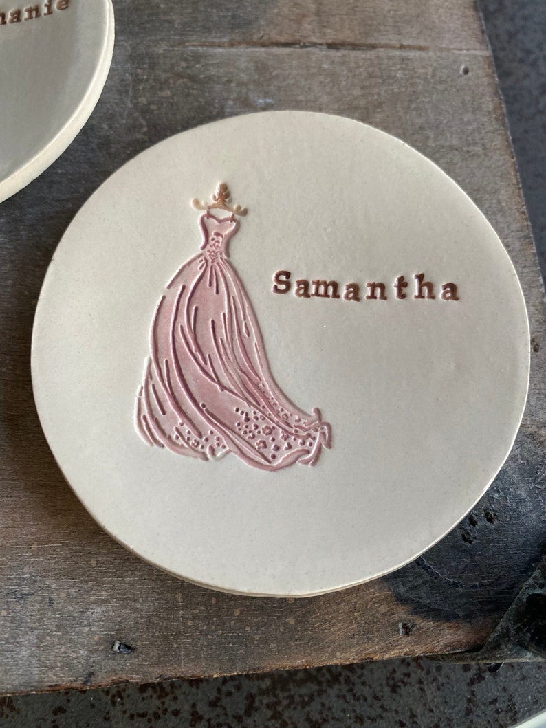 May include: A white ceramic coaster with a pink embossed design of a wedding dress and the name "Samantha" in gold lettering.