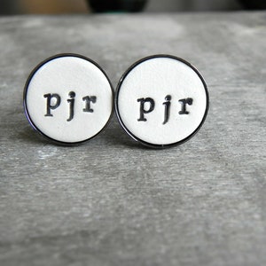 May include: Two white round cufflinks with a black metal rim. The cufflinks have the letters "pjr" printed in black on the white surface.